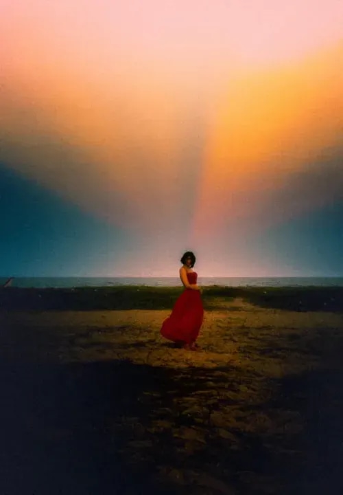A woman in a bright, red dress in the distance in a field with a colorful sky.