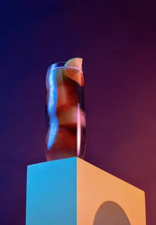 A glass of soda sitting on a pedestal in front of a vibrant purple background.