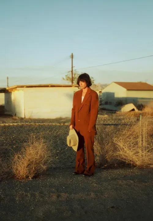 A person in a a vintage suit standing in a rural, western yard.